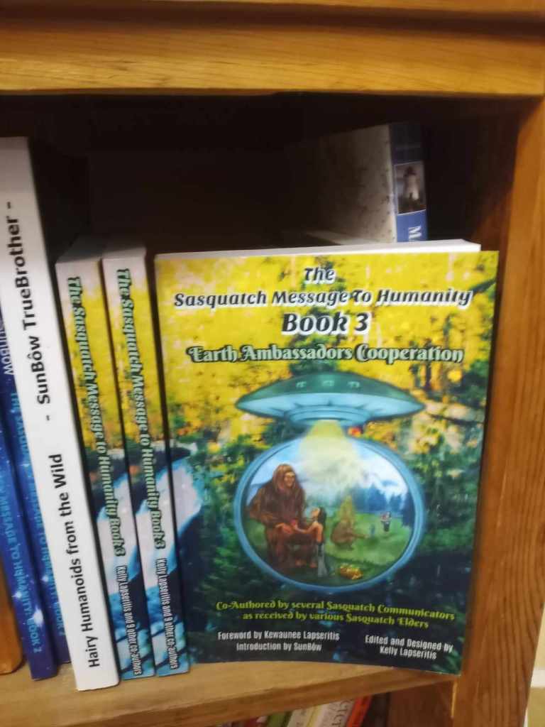 A close-up view of a bookshelf displaying several books, with the focus on 'The Sasquatch Message to Humanity Book 3: Earth Ambassadors (Cooperation)' in vibrant yellow and green cover, featuring an illustration of a Sasquatch and a humanoid figure.