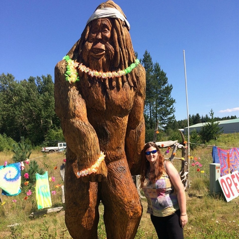 A woman stands beside a large wooden statue of Sasquatch, decorated with a floral necklace, in a colorful outdoor setting with vibrant banners in the background.